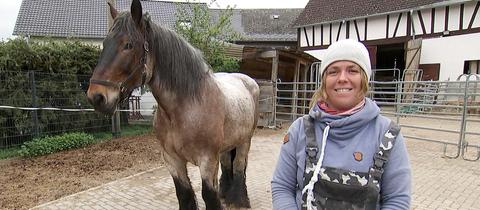 Stefanie Kirchner (im Bildvordergrund) steht im Hof und lächelt in die Kamera. Neben ihr steht ihr braun-graues Pferd.