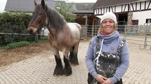 Stefanie Kirchner (im Bildvordergrund) steht im Hof und lächelt in die Kamera. Neben ihr steht ihr braun-graues Pferd.