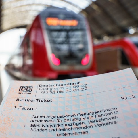 Ein 9-Euro-Ticket in Papierform - scharf im Bildvordergrund. Züge im Bahnhof unscharf im Bildhintergrund.