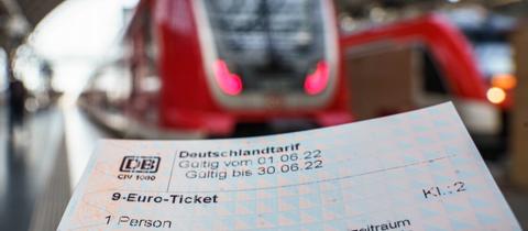 Ein 9-Euro-Ticket in Papierform - scharf im Bildvordergrund. Züge im Bahnhof unscharf im Bildhintergrund.