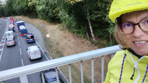 Kristina Post steht auf einer Brücke über einer Straße,auf der sich viele Autos stauen. Sie trägt eine gelbe Jacke und einen gelben Helm und blickt in die Kamera (Selfie)