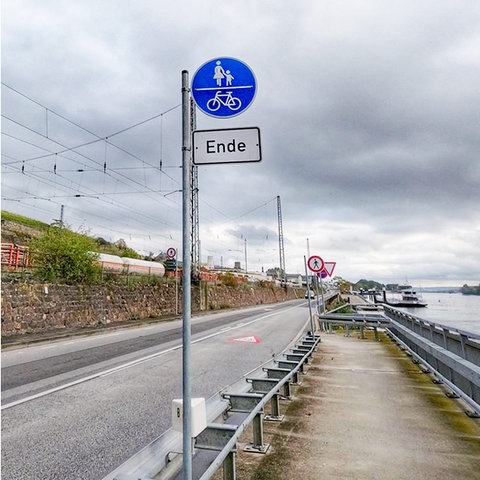 Kombo von zwei Fotos: links Radfahrer auf einem Radweg, der zwischen einer Straße und dem Fluß Rhein entlangführt; rechts ein Foto eines blauen Schildes mit Radfahrer- und Fußgänger-Symbol und darunter eine Extraschild "Ende". 