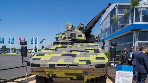 Ein grünkarierter Panzer umzäunt auf einer Ausstellungsfläche. Daneben stehen Menschen in Anzügen und ein erklärendes Schild. Im Hintergund Stangen mit "Rheinmetall"-Fahnen.