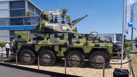 Ein grünkarierter Panzer umzäunt auf einer Ausstellungsfläche. Daneben stehen zwei Menschen und schauen. Auf der anderen Seite eine Stange mit einer "Rheinmetall"-Fahne.