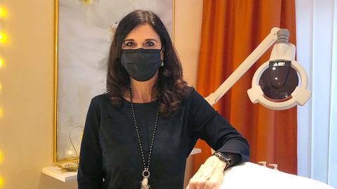 Eine Frau mit dunklen Haaren und schwarzer Maske steht vor einer Liege.