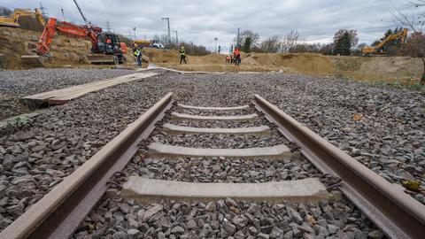 Ein Bahngleis endet in einem Haufen Schotter, dahinter sind Bagger und Bauarbeiter zu sehen.