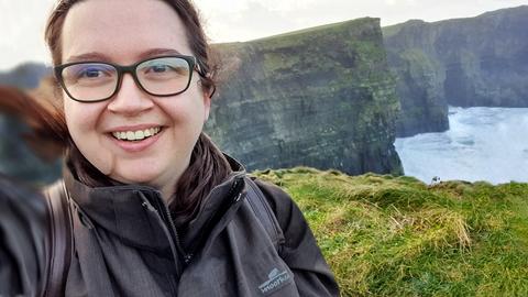 Im linken Bildvordergrund: Kopf einer lachenden jungen Frau mit Brille. Im rechten Bildhintergrund Teil einer schroffen Steilküste und Meer.