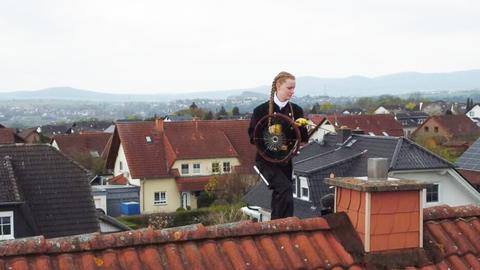 Schornsteinfegerin auf dem Dach, wie sie das Putzgerät in den Schorstein führt. Im Hintergund eine bergige Landschaft