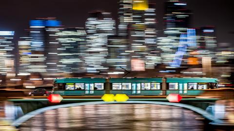 Eine Straßenbahn fährt nachts über die Ignatz-Bubis-Brücke in Frankfurt vor der erleuchteten Skyline