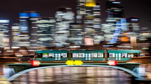 Eine Straßenbahn fährt nachts über die Ignatz-Bubis-Brücke in Frankfurt vor der erleuchteten Skyline