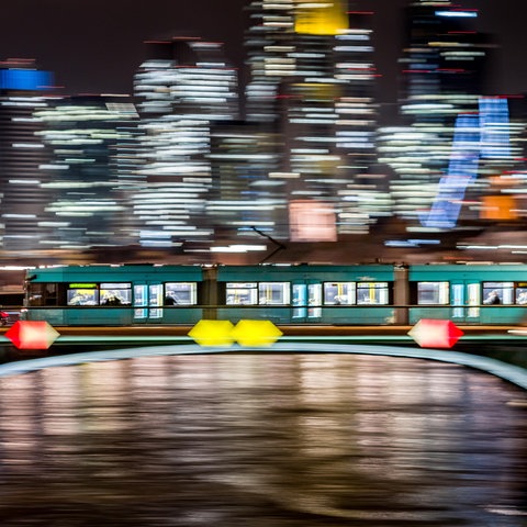 Eine Straßenbahn fährt nachts über die Ignatz-Bubis-Brücke in Frankfurt vor der erleuchteten Skyline