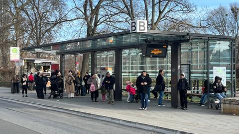 Wartende Menschenmenge an Bushaltestelle in Wiesbaden neben einem Informationsschild, auf dem steht: "Bitte auf Wagenbeschilderung und Fahrplanaushang achten"