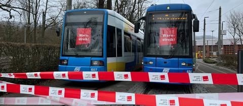 Das Bild zeigt zwei Straßenbahnen auf Gleisen. In den Scheiben der Fahrerkabinen hängen rote Schilder mit der Aufschrift "Warnstreik". Im Vordergrund sind weiß-rote Absperrbänder mit der Aufschrift "Verdi" zu sehen.