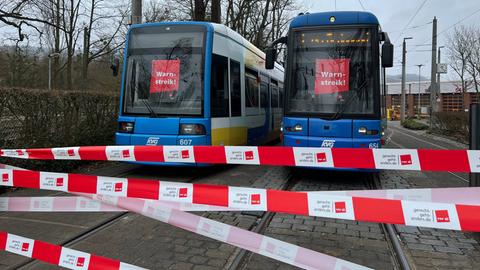 Das Bild zeigt zwei Straßenbahnen auf Gleisen. In den Scheiben der Fahrerkabinen hängen rote Schilder mit der Aufschrift "Warnstreik". Im Vordergrund sind weiß-rote Absperrbänder mit der Aufschrift "Verdi" zu sehen.