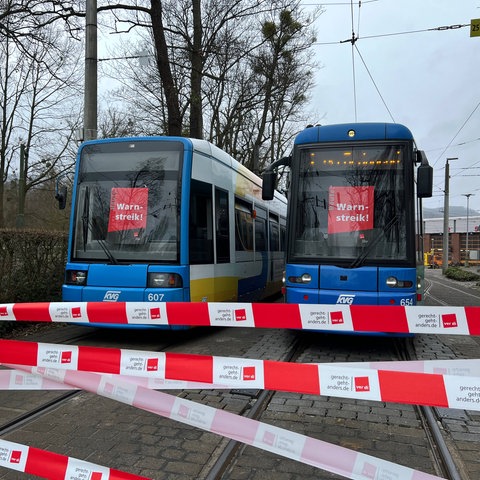 Das Bild zeigt zwei Straßenbahnen auf Gleisen. In den Scheiben der Fahrerkabinen hängen rote Schilder mit der Aufschrift "Warnstreik". Im Vordergrund sind weiß-rote Absperrbänder mit der Aufschrift "Verdi" zu sehen.