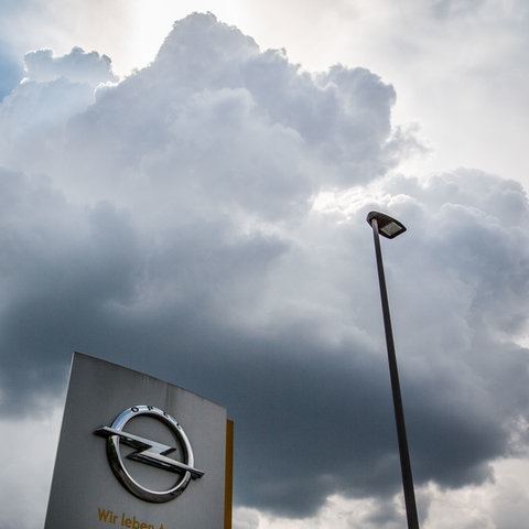 Opel-Symbol auf einer Stele vor dem Werk in Rüsselsheim - aus der Froschperspektive fotografiert. Darüber schwebt eine dunkle Wolke am Himmel.