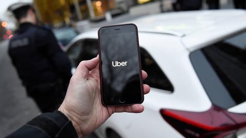 Stadtpolizei kontrolliert Uber-Fahrer: Ein Polizist steht auf der Straße neben einem Uber-Auto, im Vordergrund eine Hand mit einem Handy, auf dessem Display "Uber" steht.