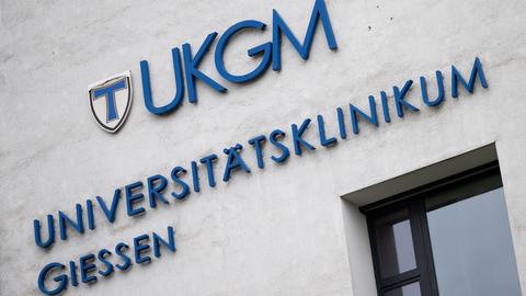 Blauer Schriftzug "UKGM Universitätsklinikum Gießen" an einer grauen Hauswand.