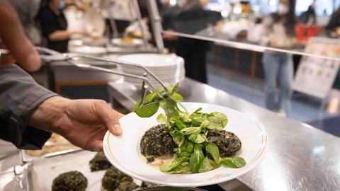 Großaufnahme eines Tellers in einer Großküche, auf dem Knödel angerichtet sind. Jemand legt mit einer Zange Feldsalat auf den Teller.