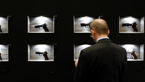 Ein Mann steht vor einem Regal in dem hinter Sicherheitsglas einzelne Pistolen ausgestellt sind. 