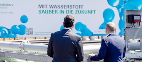 "Mit Wasserstoff sauber in die Zukunft" steht auf einem Banner im Außenbereich. Davor stehen zwei Männer mit dem Rücken zur Kamera.