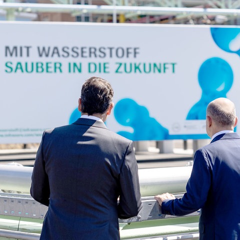 "Mit Wasserstoff sauber in die Zukunft" steht auf einem Banner im Außenbereich. Davor stehen zwei Männer mit dem Rücken zur Kamera.