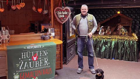 Ein Mann mit grauen Haaren steht vor weihnachtlich geschmückten Buden. Er hält einen Rauhaardackel an einer Leine.